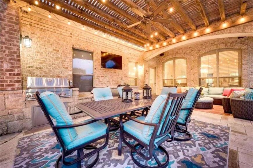 A closer view shows the summer kitchen, metal dining table, and wicker seats topped with light blue cushions and multi-colored pillows.