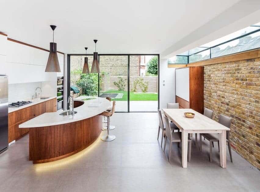 Layout: An exceptionally stylish kitchen space with dining area is made remarkable through several features. A brick facade on the back wall brings a rustic charm, while white countertops are the perfect toppers for impeccable craftsmanship with the wooden cabinetry. The room itself is so open and airy thanks to the entirely windowed wall and skylights. Cement tiles are a popular choice for more modern kitchen designs, but everything all put together still feels very warm and inviting. Dining Set: A super lightly stained birch wood table is long and narrow, making it the perfect table to host dinners with plenty of guests and also plenty of elbow room. I personally would have chosen a different set of chairs (maybe with a midcentury modern inspiration) to carry on with the wooden features of the room. Decoration: Here we see a wonderfully simple centrepiece: an artisanal wooden bowl filled with some fresh fruit. Though the effect isn't really felt from so far away, a centrepiece as simple as this is really what this type of room demands. A still life! This perfect dining and kitchen area was created by Inspired Dwellings