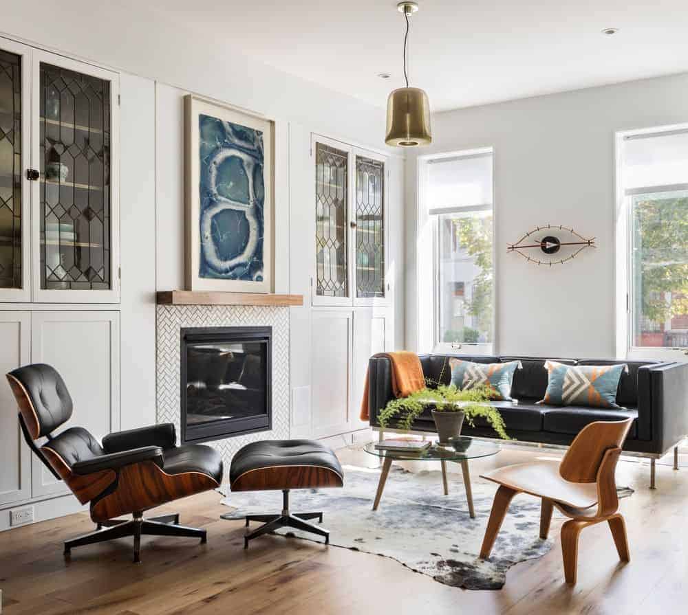 This living room with black couch and glass circular table on the rug. It also has a fireplace and white walls with decors.