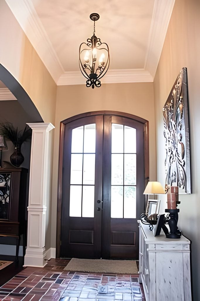The foyer has a wooden french front door, an ornate pendant and a distressed console table under the decorative artwork.