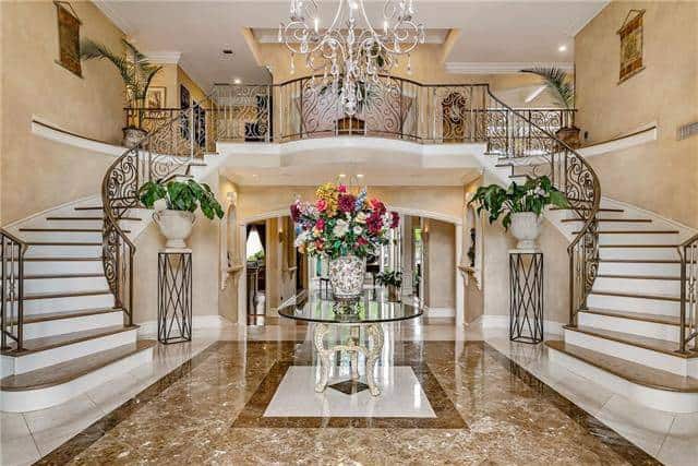 The foyer is filled with a round glass top center table, ornate chandelier, and flower pedestals sitting against the bifurcated staircase.