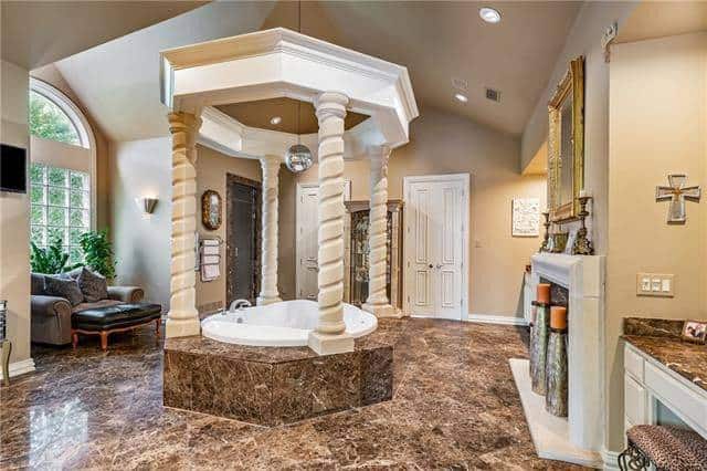 Primary bathroom with granite top vanities, a fireplace, gray couch, and an elegant deep soaking tub surrounded by spiral columns.