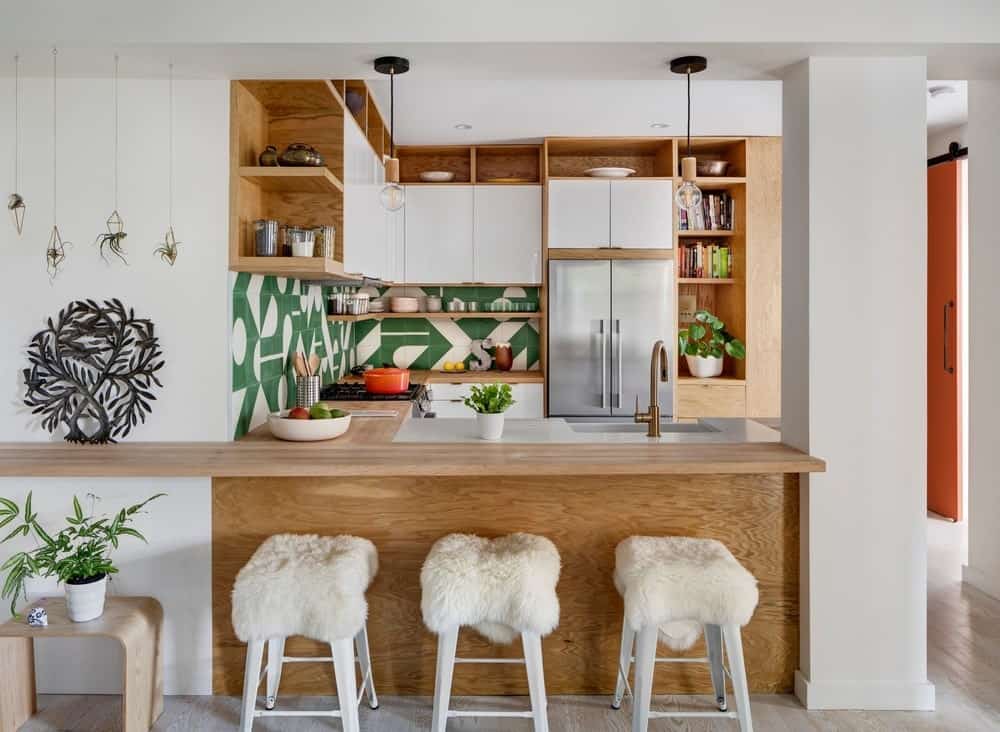 This view of the kitchen showcases the L-shaped peninsula with a wooden breakfast bar paired with white stools to match the white walls. This peninsula is attached to a thick white column that connects the floor to the white ceiling.