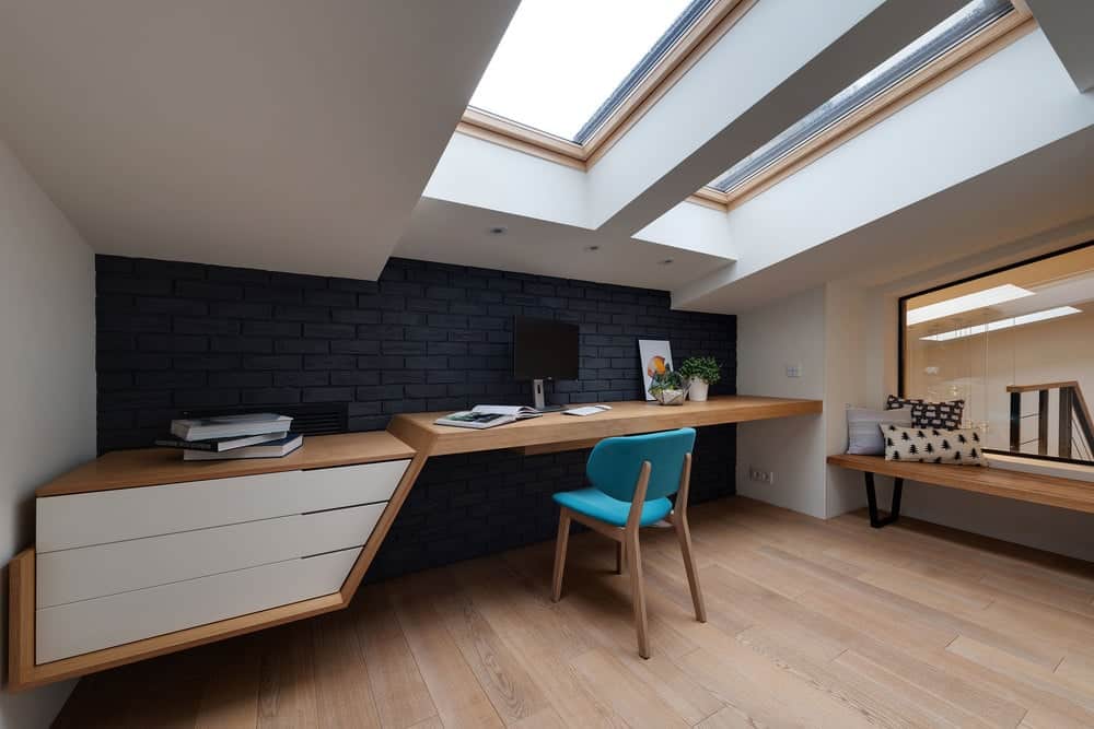 The hardwood flooring matches with the surface of the built-in desk that has white drawers to match the white shed ceiling with skylights. This is then contraste dby the black brick wall of the desk.