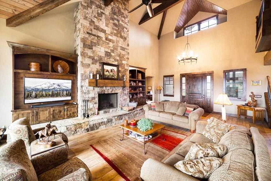 The living room has comfy beige seats and a stone fireplace flanked by the TV and the wooden bookcase.