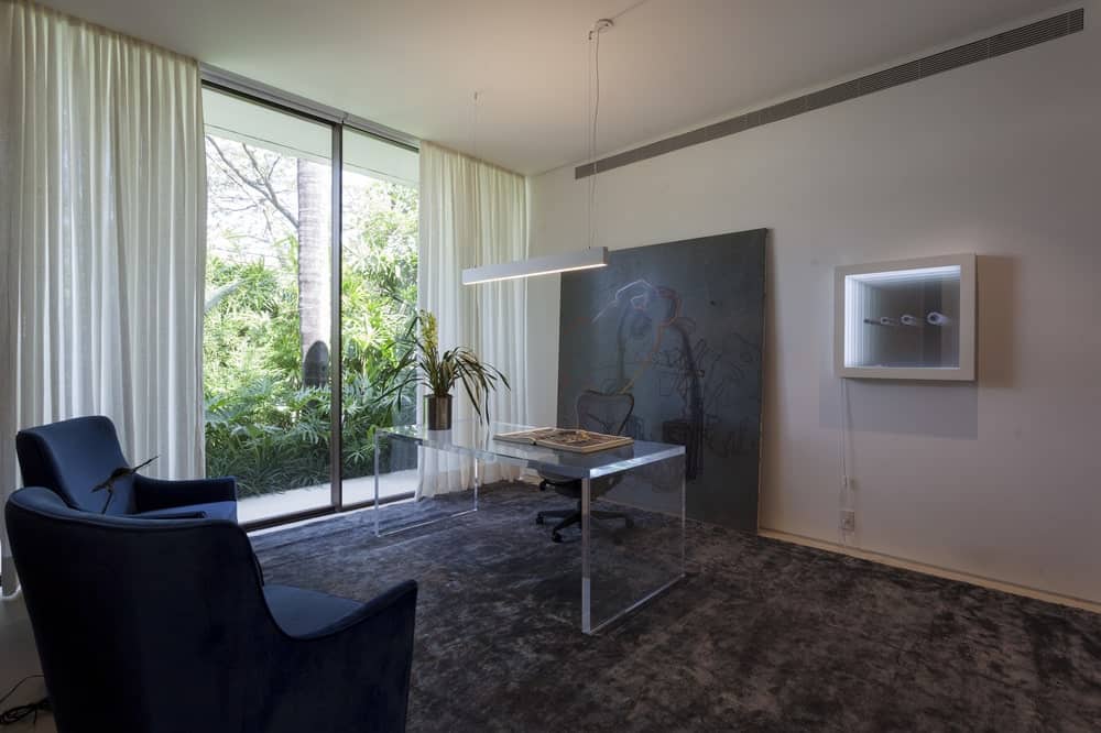 This is a sophisticated modern home with a transparent desk. This pairs quite well with the black carpeting and black wall artwork behind the black swivel chair that stands out against the white walls and ceiling.
