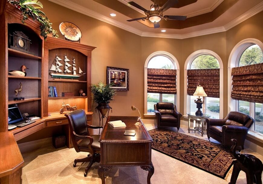 The study has wooden furnishings, a stunning tray ceiling, and arched windows dressed in patterned shades.