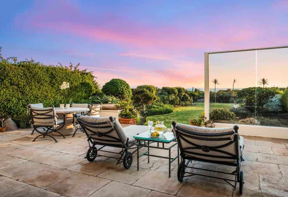 This is the patio of the main level that has a couple of comfortable cushioned lawn chairs flanking a glass-top side table. Beside this is the outdoor dining area with a round dining table with a background of tall shrubs. These are on beige tiles that transitions to the backyard grass lawn.