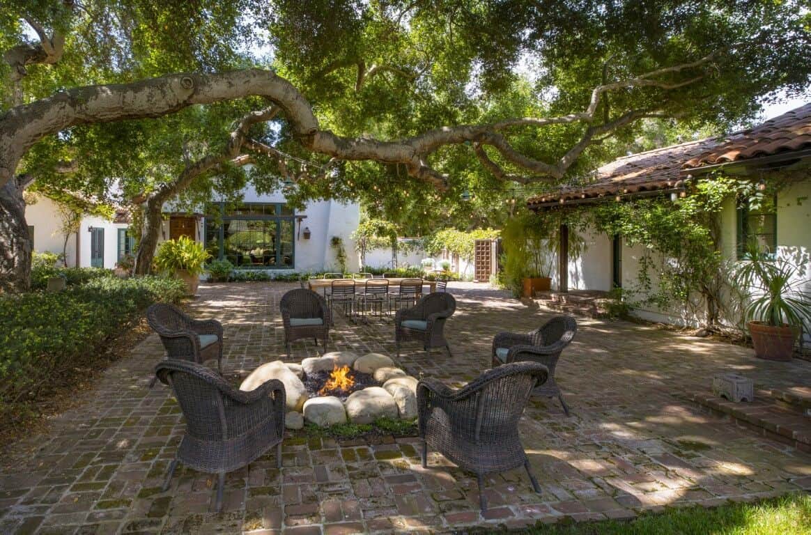 This is the charming backyard patio with a set of outdoor armchairs surrounding the rustic firepit in the middle of the brick flooring under the shade of a big tree.