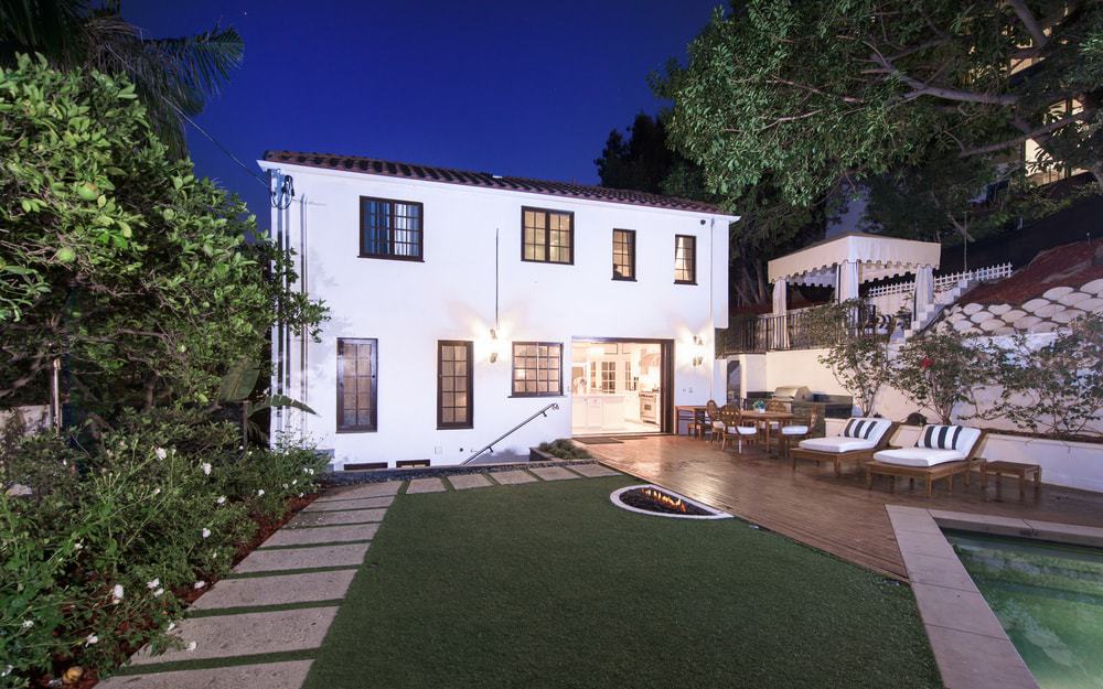 This is the beautiful backyard patio of the house with a wooden deck flooring fitted with various sitting areas. There is an attached half-circle firepit to this as well as a concrete slab walkway.