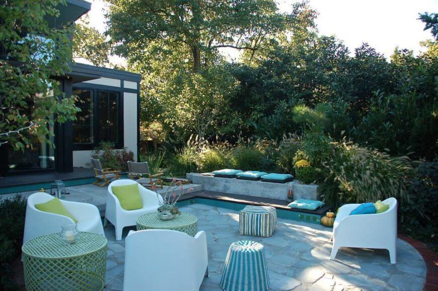 The back patio shares a large group of novel seating alternatives, with white easy chairs and wicker tables tucked away in rich greenery behind the home.