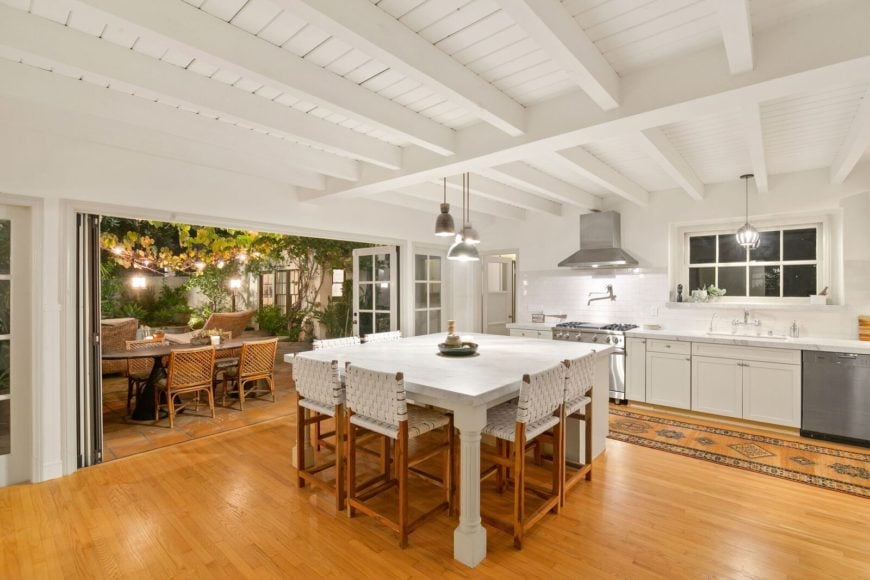 This is a bright and white kitchen with a white beamed ceiling and a large white kitchen island with a built-in table paired with wooden chairs to match the hardwood flooring.