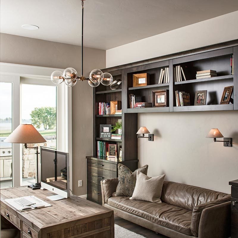 The study has a glass chandelier, a natural wood desk, and a brown leather sofa lit by warm sconces.