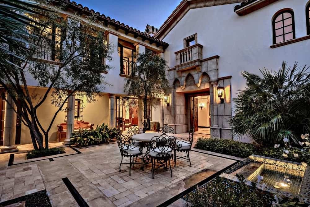 This is a luxurious outdoor patio with a beige marble flooring edged and accented with contrasting black granite complemented by the surrounding shrubs and trees as well as a small fountain.