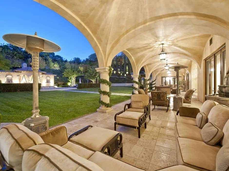 This is a large patio with sitting areas underneath a row of groin vault ceiling with pillars that match well with the beige flooring tiles. These stop at the edge of the grass lawn that leads to the backyard.