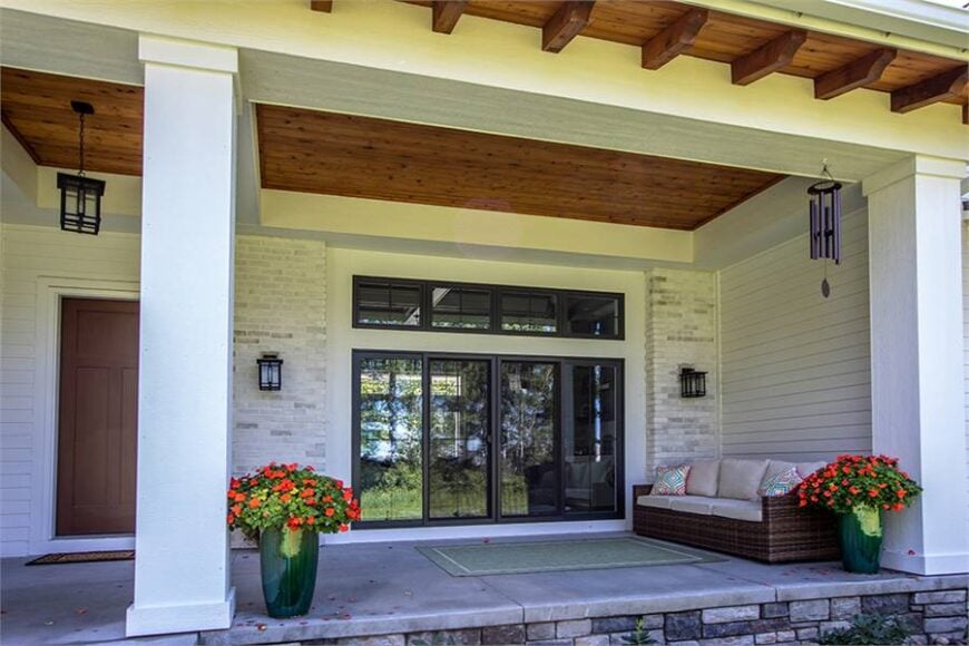 The covered front porch is filled with lovely potted plants, a wicker cushioned sofa, and a bordered rug that lays on the concrete flooring.