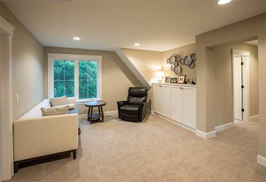 The bonus room is filled with a beige sofa, a small round table, a black leather recliner, and a white built-in cabinet topped with framed photos.