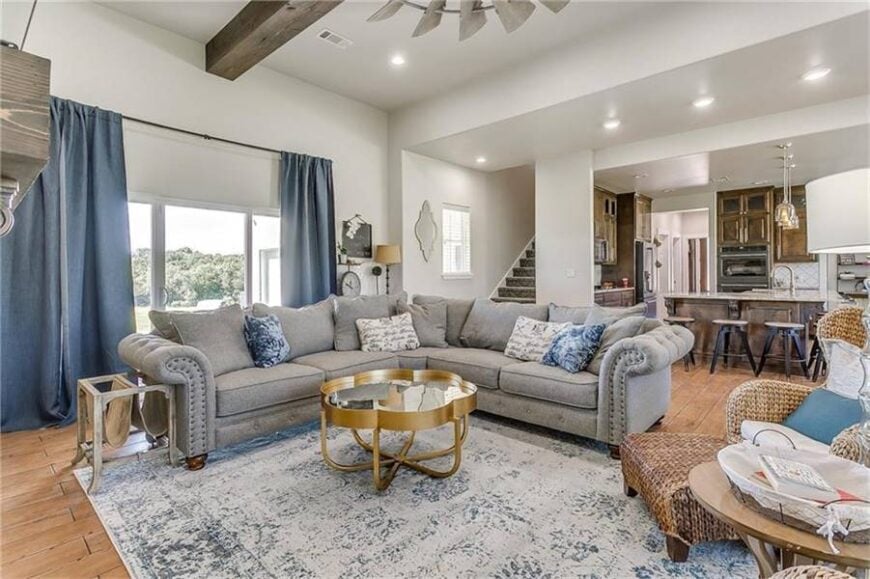 Across the kitchen is the family room with a tufted V-shaped sectional, glass top coffee table, and a wicker lounger over a distressed area rug.