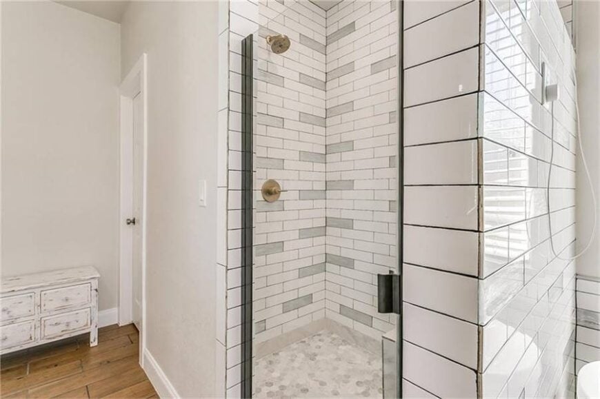 A closeup view of the walk-in shower with hex tile flooring and white brick walls mounted with brass fixtures.