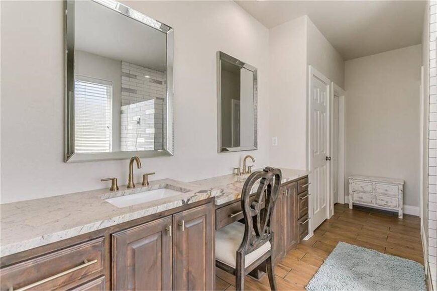 Multi-level vanity with granite countertops and undermount sinks. It is paired with chrome framed mirrors and a cushioned chair.