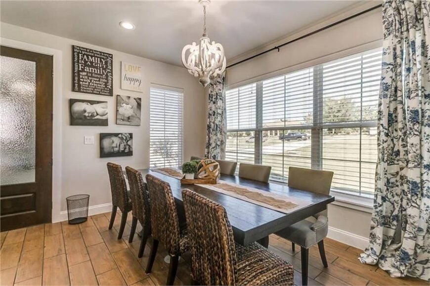 The dining room offers wicker and gray upholstered chairs along with a dark wood dining table lit by an ornate chandelier.