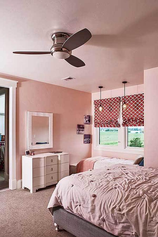 This bedroom has light pink walls, gray carpet flooring, and white framed windows dressed in red patterned roman shades.