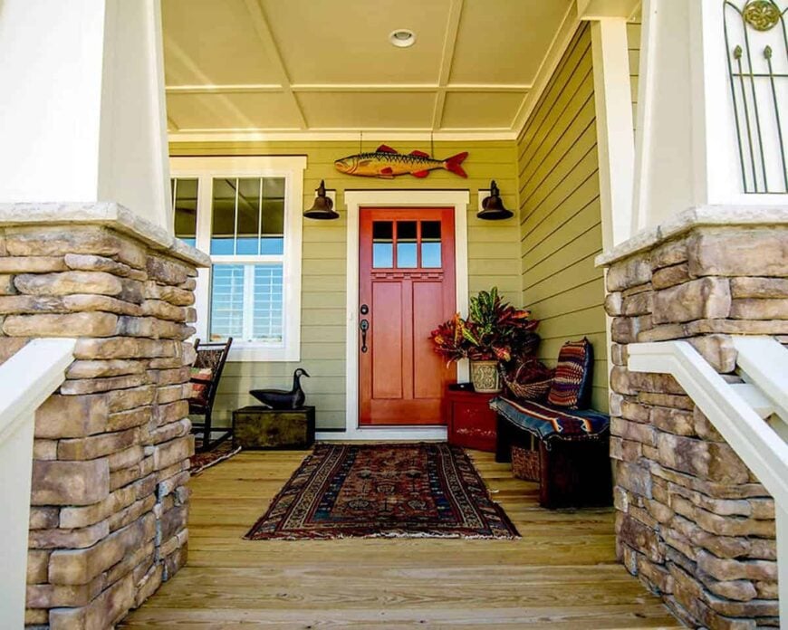 Covered front porch with cozy seats, tribal rugs, and a red front door fixed against the green shiplap walls.
