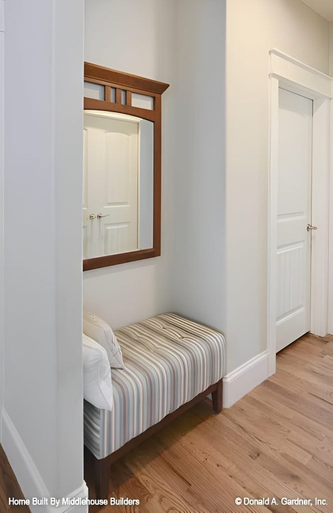 This nook has a hardwood flooring, a wooden framed mirror, and a striped tufted bench topped with white pillows.