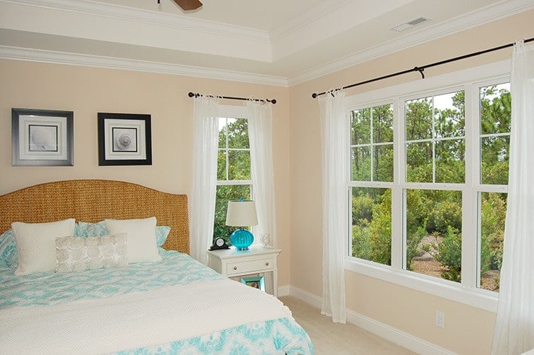 The primary bedroom has a cozy wicker bed and white framed windows dressed in sheer curtains.