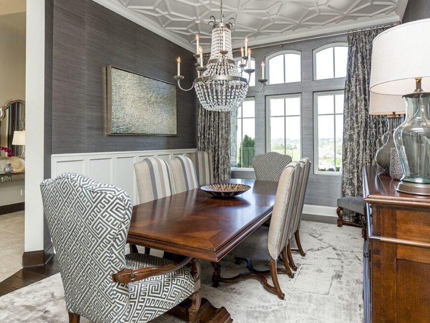 A beaded chandelier hanging from the stylish tray ceiling illuminates the dining room that offers a wooden buffet bar and rectangular dining set.