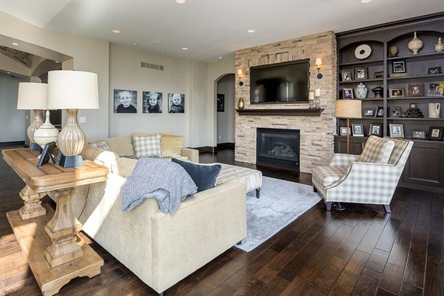 Hearth room with a stone fireplace, dark wood built-ins, an L-shaped sofa, and a checkered lounge chair that matches the fluffy pillows.
