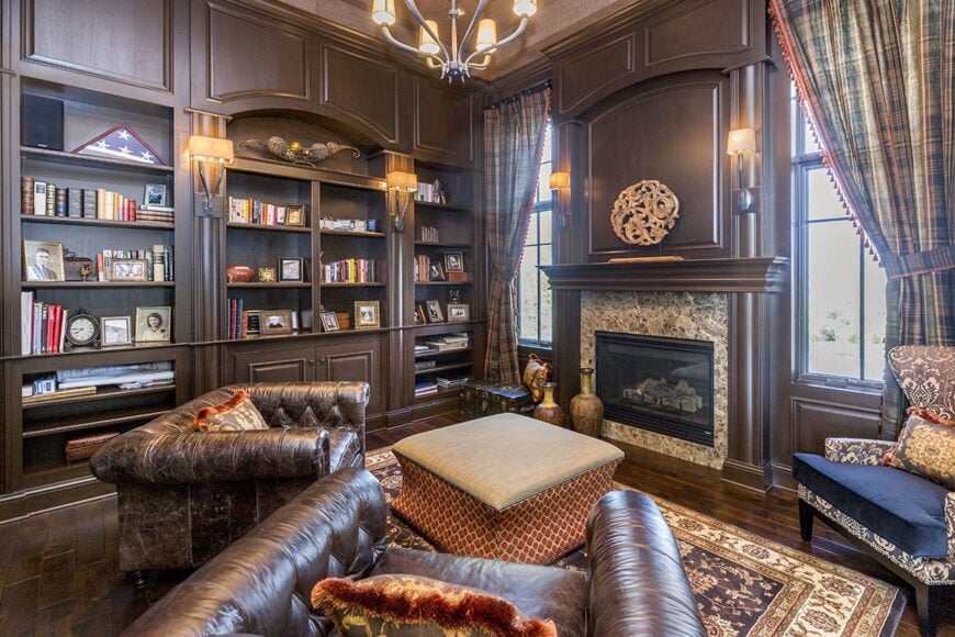 The study offers paneled built-ins, a wingback and leather tufted chairs, and a patterned ottoman facing the glass-enclosed fireplace.