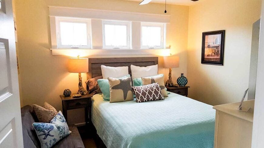 The primary bedroom has white framed windows, a cozy gray sit, and an upholstered bed flanked by wooden nightstands and drum table lamps.