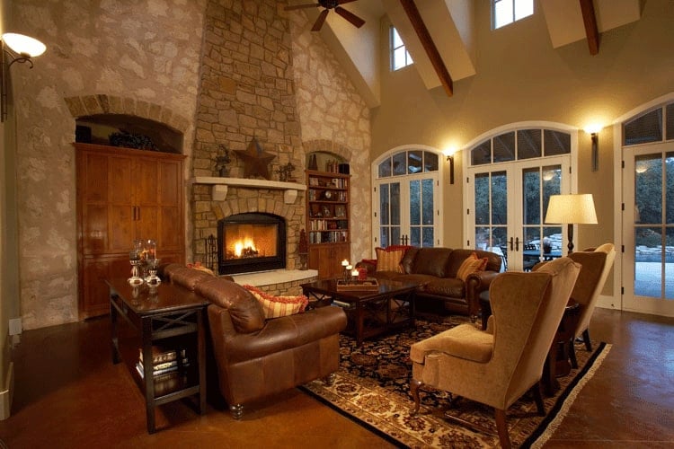 The living room offers brown leather sofas, beige wingback chairs, and a wooden coffee table facing the warm fireplace.