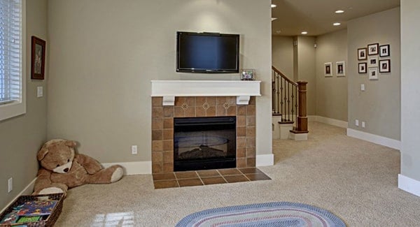 The recreation room offers a wall-mounted TV, a tiled fireplace, and an oval rug that lays on the beige carpet flooring.