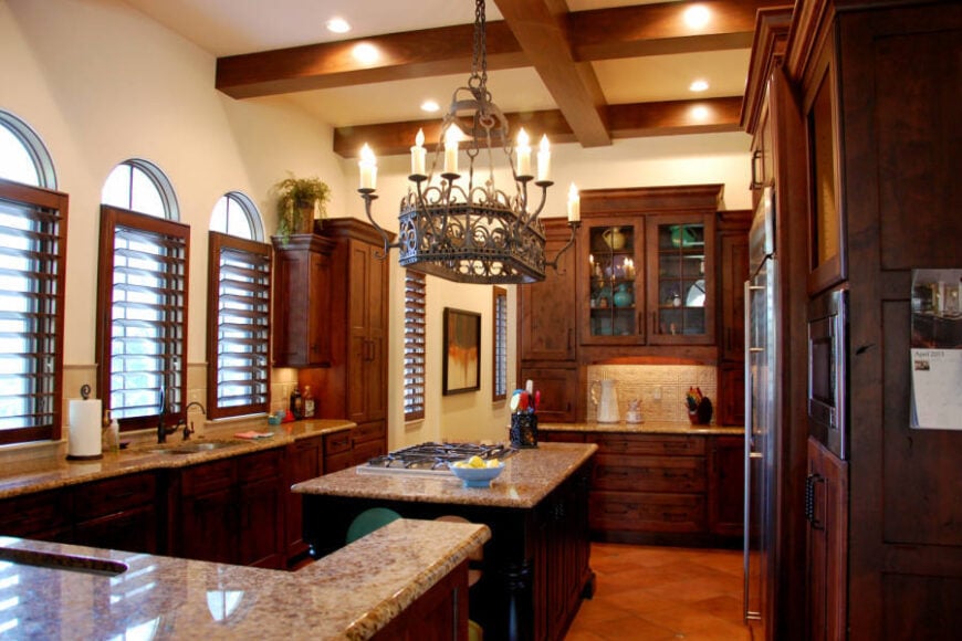 The kitchen has arched louvered windows and a beamed ceiling mounted with an ornate candle chandelier that's suspended above the center island.