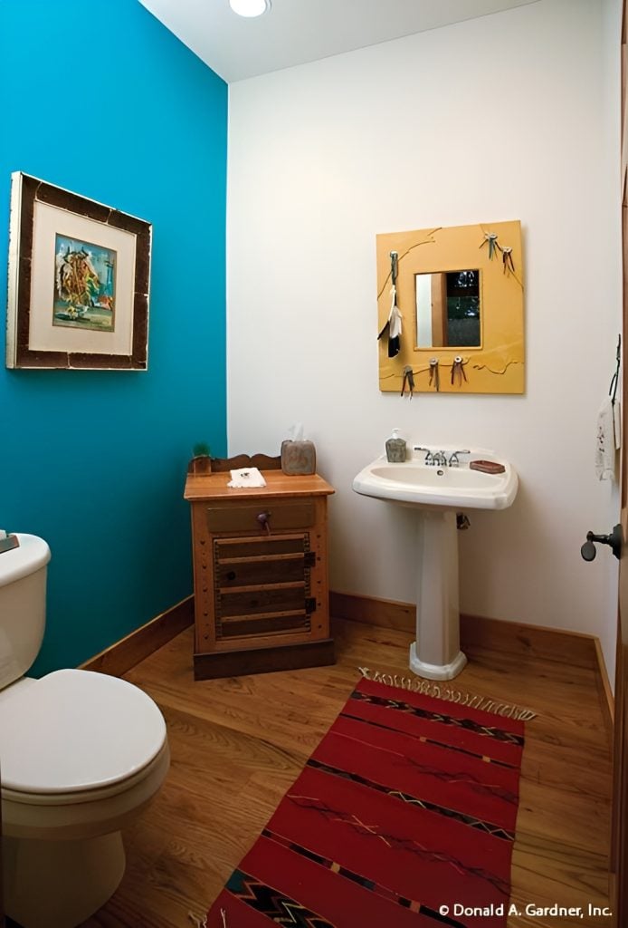 This bathroom offers a toilet, a wooden cabinet, a pedestal sink under the framed mirror, and a red tasseled runner that lays on the hardwood flooring.