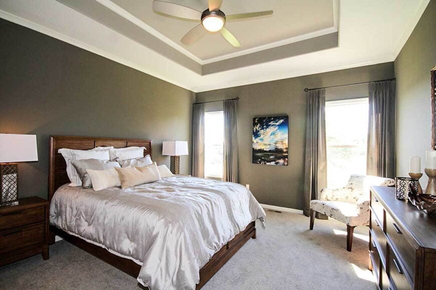 Primary bedroom with dark wood furnishings, beige carpet flooring, and a striking tray ceiling mounted with a sleek ceiling fan.