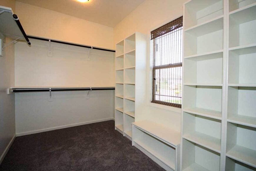 Walk-in closet with brown carpet flooring, white built-in shelves, and a glass window covered in a sheer wicker shade.