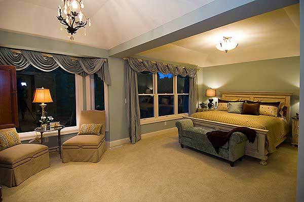 The primary bedroom offers a cozy wooden bed with a velvet bench at its foot, beige skirted chairs, and large white framed windows adorned with classy valances.The primary bedroom offers a cozy wooden bed with a velvet bench at its foot, beige skirted chairs, and large white framed windows adorned with classy valances.