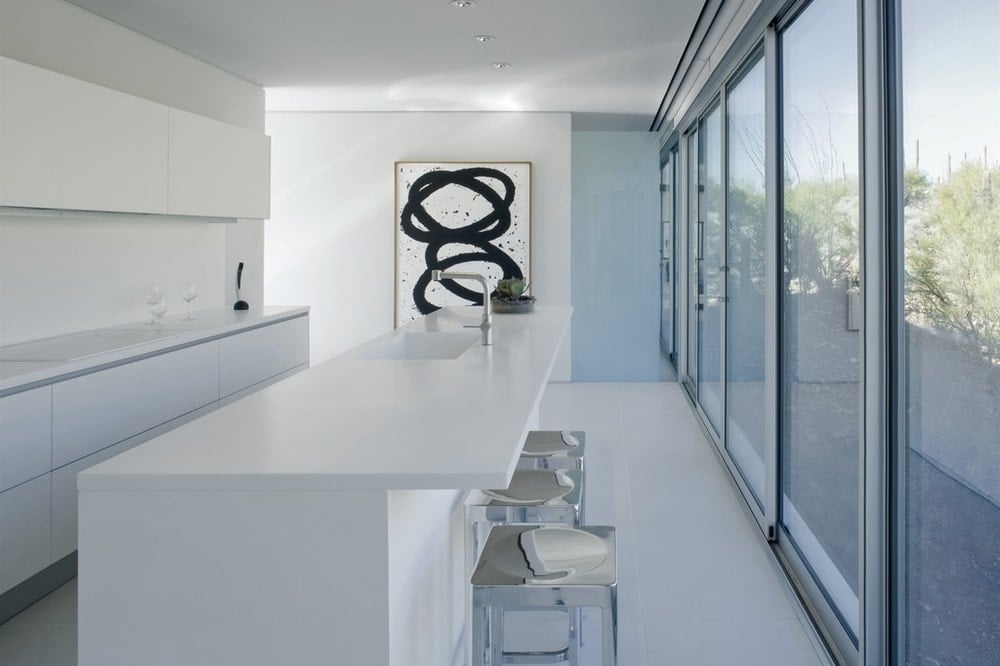 This other view of the kitchen showcases the stainless steel stools paired with the long white breakfast bar adorned with a black and white painting on the far wall. Image courtesy of Toptenrealestatedeals.com.