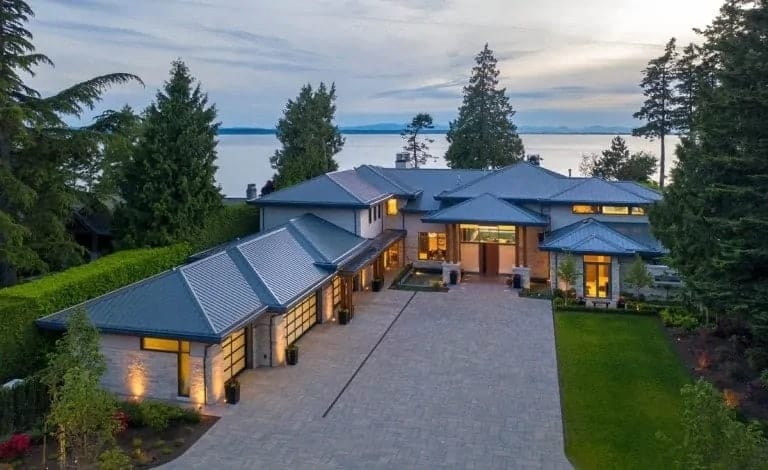 This is an aerial view of the house showcasing the wide concrete driveway and the L-shaped house a warm glow and earthy tone to its walls that pair well with the surrounding landscape.