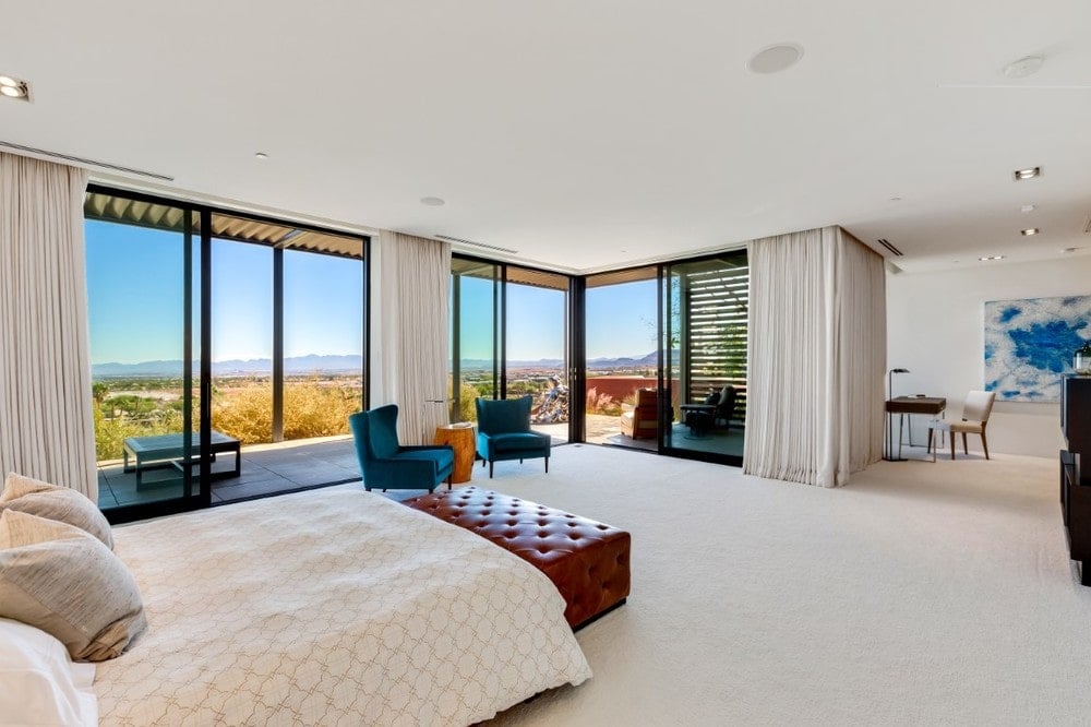 This is a spacious bedroom with a bed that blends with the beige walls, ceiling and floor. These are then complemented by the red leather cushioned bench at the foot of the bed and the pair of green cushioned chairs by the glass walls. Image courtesy of Toptenrealestatedeals.com.