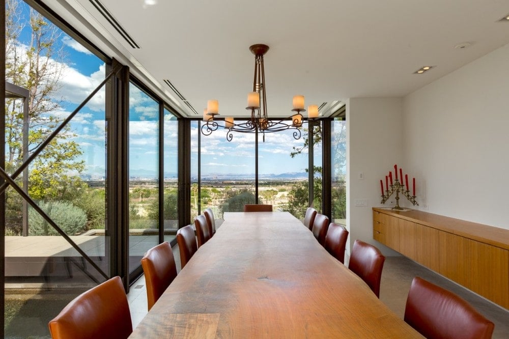 This is a close look at the dining room with a large rectangular wooden dining table surrounded by red leather chairs, topped with a chandelier and brightened by the glass walls. Image courtesy of Toptenrealestatedeals.com.