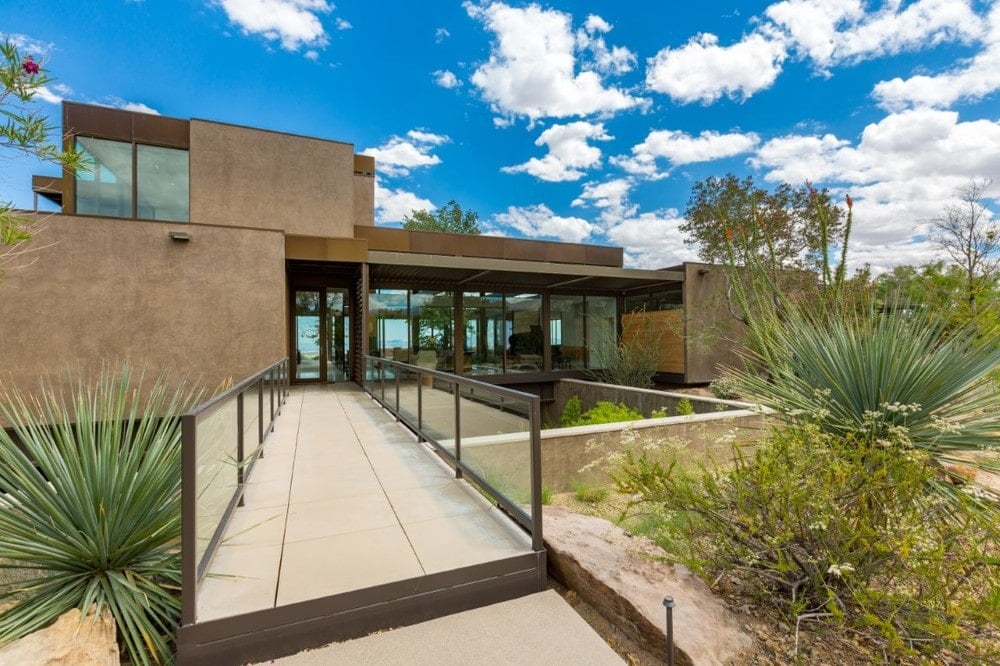 This is a look at the front of the house featuring the main entrance with concrete walkway bordered with glass to match the glass walls and glass doors of the house that stands out against the earthy tone of the exteriors. Image courtesy of Toptenrealestatedeals.com.