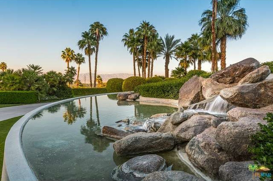 This is a close look at the landscaping with a large stone waterfall water display and a background of tall palm trees. Image courtesy of Toptenrealestatedeals.com.