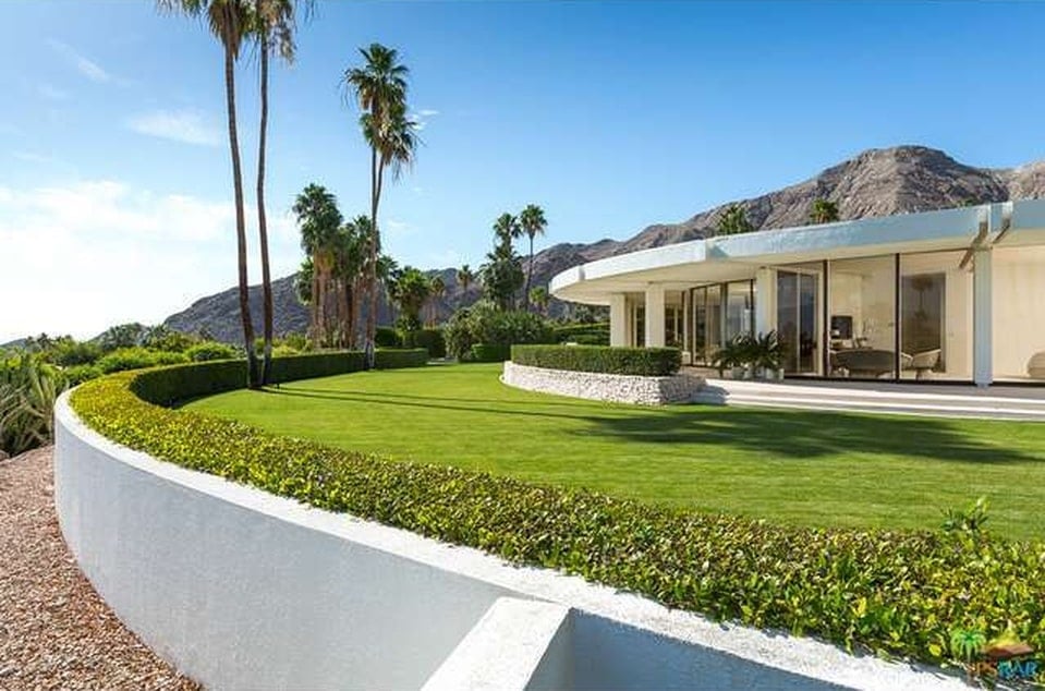 This is a look at the well-maintained grass lawn at the side of the house adorned with a hedge of shrubs and tall palm trees. These green elements pair well with the bright exteriors of the house. Image courtesy of Toptenrealestatedeals.com.