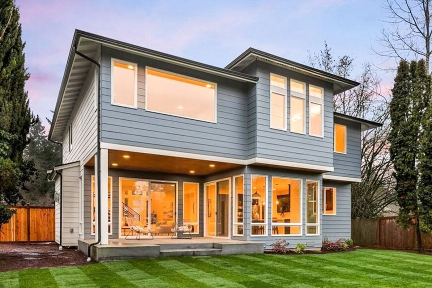 View of the house from the backyard showing its lap siding and glazed windows flooded with warm, ambient lights.