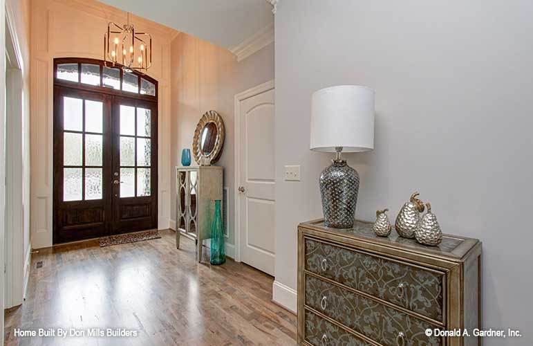 The foyer has a wooden french door, candle chandelier, and a stylish console table topped with a round mirror and transparent blue vase.