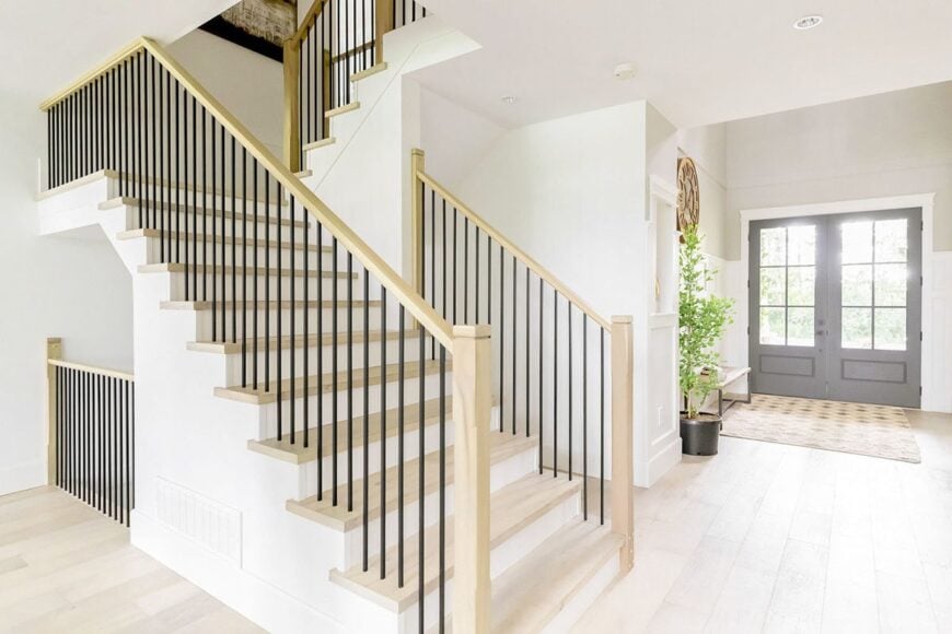The foyer is filled with a staircase, french entry door, a wooden bench, fresh potted plant, and a patterned rug that lays on the wide plank flooring.